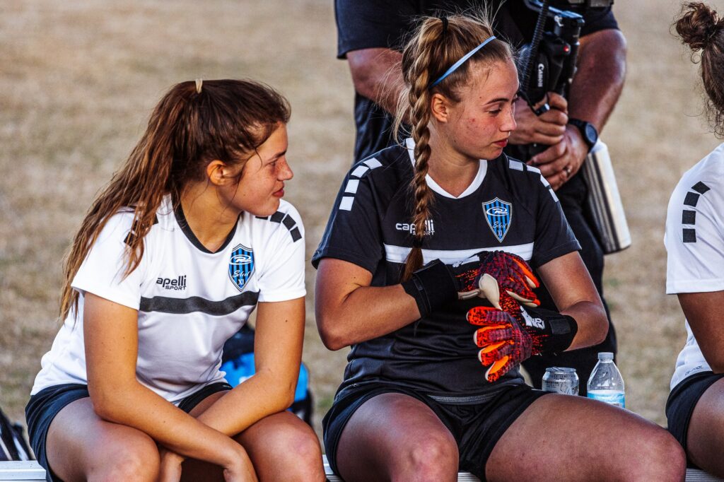 Two female soccer players sit on a bench, one in a white jersey and the other in a dark gray jersey with gloves, appearing to have a conversation. Both wear shorts with an emblem on their shirts. The background features blurred figures and equipment.