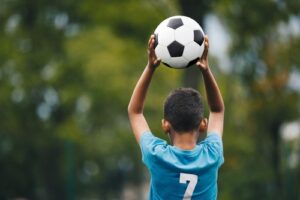 A young soccer player, viewed from the back, holds a soccer ball above his head 