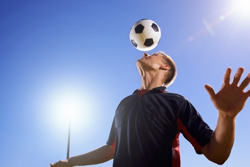 A soccer player balances a ball on his head as he practices heading on a sunny day against a clear blue sky