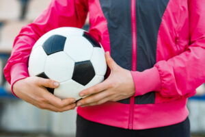 The body of a female soccer player who wears a pink and black jacket and holds a soccer ball to her side 