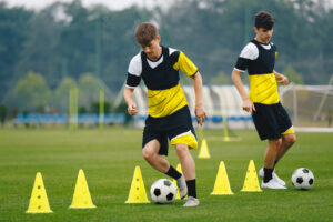 Two young male soccer players in yellow and black sports uniforms train on a grassy field. One dribbles a soccer ball through yellow cones while the other observes. A goalpost and more training equipment are visible in the background.