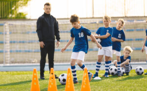 A coach looks on as a youth soccer team performs soccer practice warm-up drills with balls and cones 