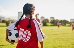 A young girl holds a soccer ball and looks out on the field and experiences the mental benefits of playing soccer