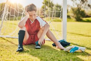 A young soccer player sits on the ground in front of the goal with one shoe off, looking defeated 