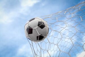 A soccer ball kicked by a member of the U.S. Women’s National Soccer Team flies into the net against a blue sky 