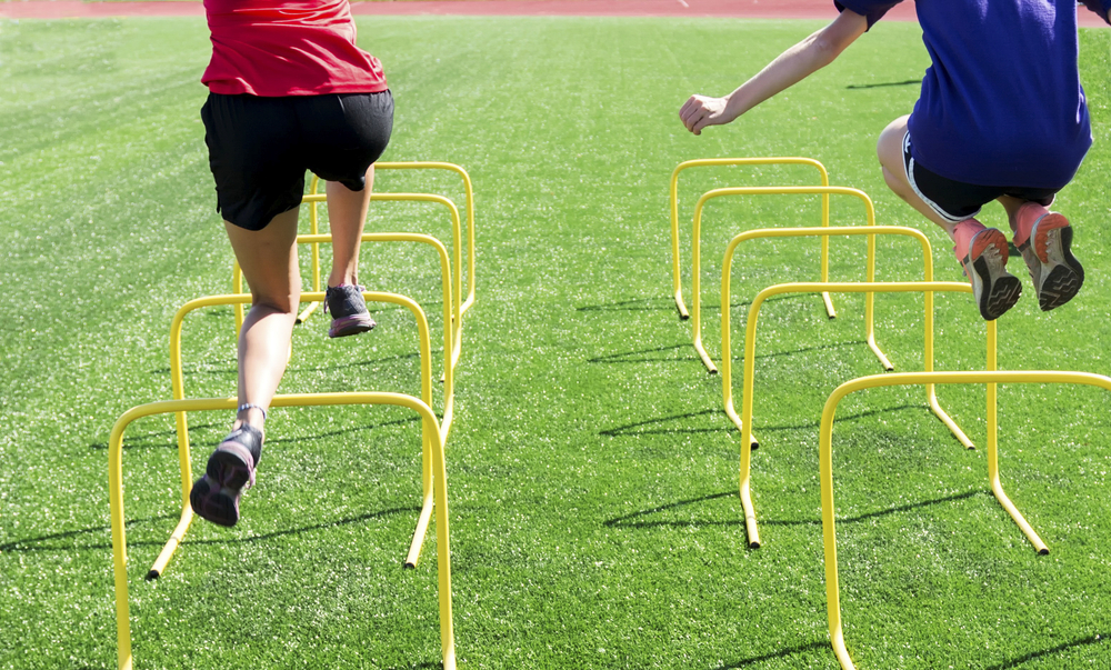 Two people are mid-air, jumping over yellow hurdles on a green field. They wear athletic shorts and shirts, with visible legs and shoes. The focus is on their action and movement in a straight line over the hurdles.