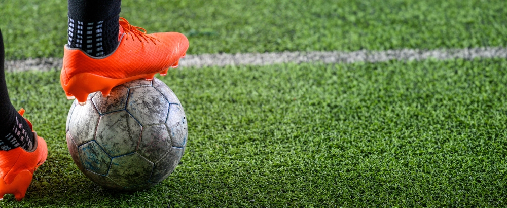 A person wearing bright orange soccer cleats stands on a worn soccer ball, positioned on a green, grassy field.